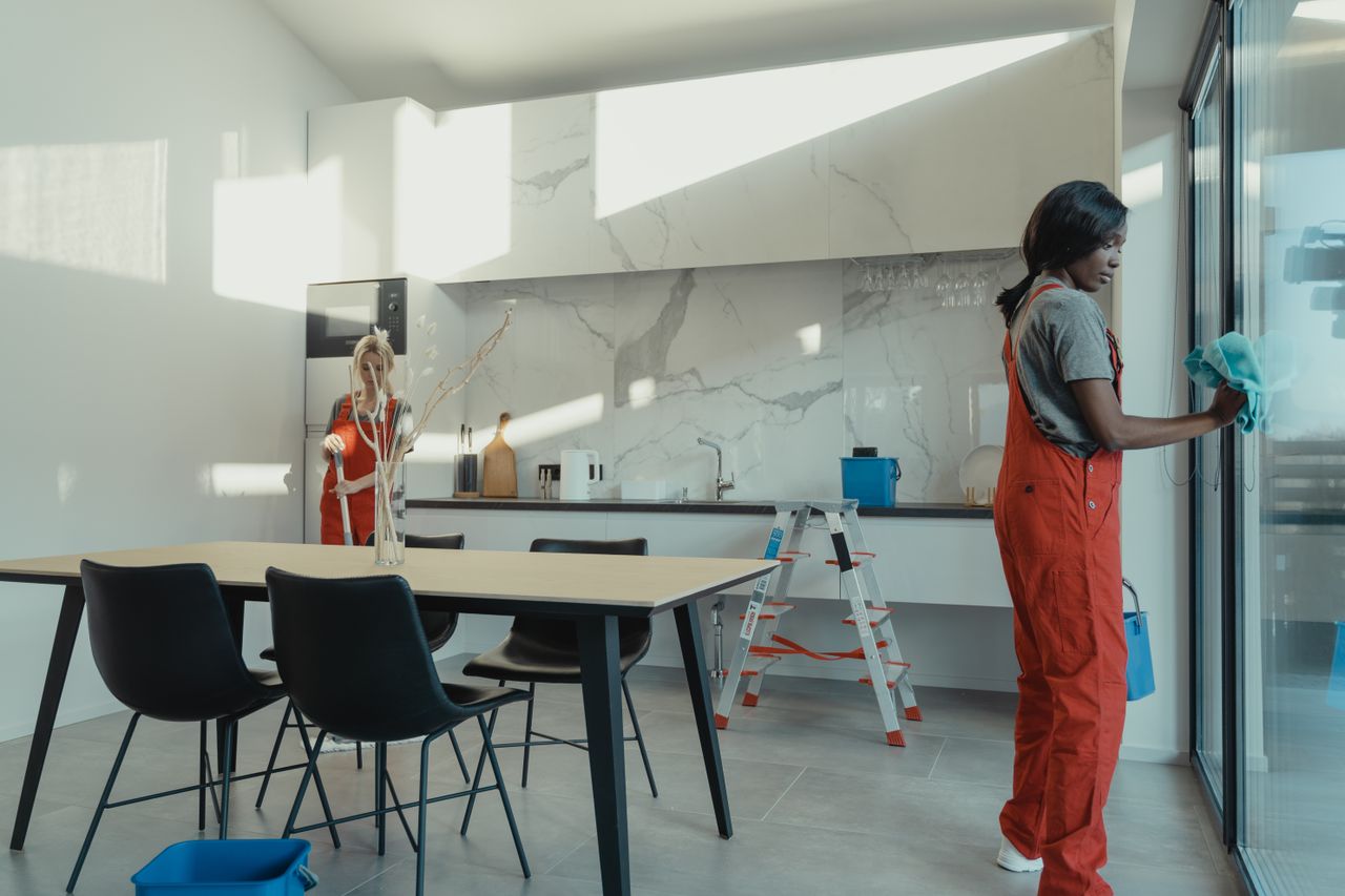 team-02 Two women in work clothes cleaning a kitchen with glass doors.